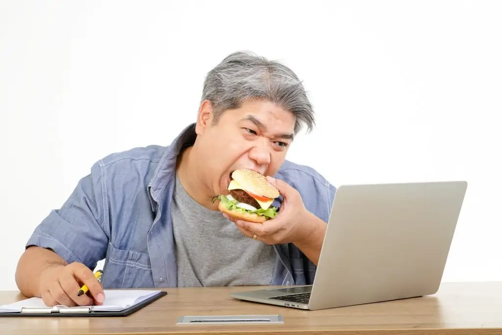 Photo of overweight man eating a burger at his desk.